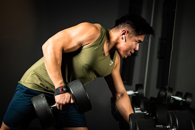 A person is engaged in a workout session, holding a dumbbell with one hand while leaning on a bench. They are wearing a green sleeveless shirt and dark shorts, with earbuds in their ears. The setting appears to be a gym, with a focused and determined expression on their face.
