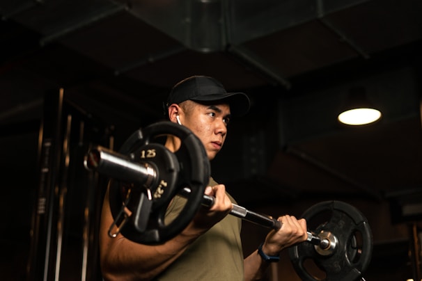 A person is engaged in a weightlifting exercise, holding a barbell with focus and determination. The setting appears to be a dimly lit gym, with industrial elements visible in the background. The individual wears a cap and earphones, suggesting a personal workout session.