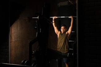 A person lifting a barbell above their head in a dimly lit gym. The room has dark walls and exercise equipment is visible, contributing to a focused atmosphere.
