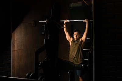 A person lifting a barbell above their head in a dimly lit gym. The room has dark walls and exercise equipment is visible, contributing to a focused atmosphere.
