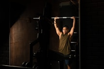 A person lifting a barbell above their head in a dimly lit gym. The room has dark walls and exercise equipment is visible, contributing to a focused atmosphere.