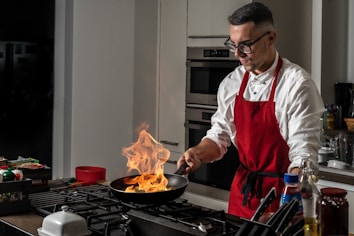 A man wearing a red apron and glasses is cooking in a modern kitchen. He is holding a frying pan over a stove, and flames are rising from the pan. The kitchen has a sleek design with stainless steel ovens in the background, and there are various kitchen items such as bottles and containers on the counter.