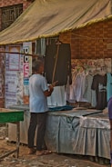 A person is standing in front of a market stall that sells clothing, looking at a black garment. The stall has a canopy for shade, and several posters and notices are displayed on the brick wall behind it. Various shirts hang on display, including a black and white checkered shirt and some white shirts.