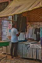 A person is standing in front of a market stall that sells clothing, looking at a black garment. The stall has a canopy for shade, and several posters and notices are displayed on the brick wall behind it. Various shirts hang on display, including a black and white checkered shirt and some white shirts.