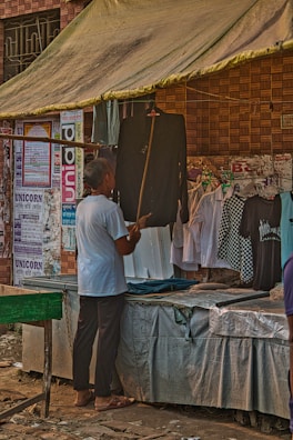A person is standing in front of a market stall that sells clothing, looking at a black garment. The stall has a canopy for shade, and several posters and notices are displayed on the brick wall behind it. Various shirts hang on display, including a black and white checkered shirt and some white shirts.