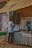 A person is standing in front of a market stall that sells clothing, looking at a black garment. The stall has a canopy for shade, and several posters and notices are displayed on the brick wall behind it. Various shirts hang on display, including a black and white checkered shirt and some white shirts.