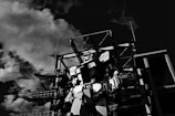 A large robotic figure stands in front of an industrial structure with metal frameworks. Clouds fill the sky, casting dramatic shadows, creating a dynamic contrast with the robot and the structure.