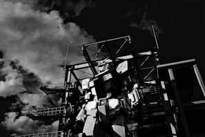 A large robotic figure stands in front of an industrial structure with metal frameworks. Clouds fill the sky, casting dramatic shadows, creating a dynamic contrast with the robot and the structure.