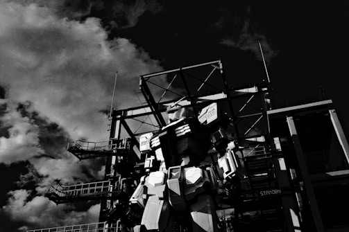 A large robotic figure stands in front of an industrial structure with metal frameworks. Clouds fill the sky, casting dramatic shadows, creating a dynamic contrast with the robot and the structure.