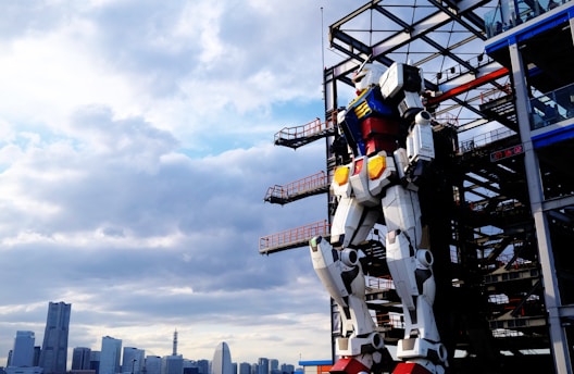 A large humanoid robot stands against a metal framework structure, overlooking a cityscape with tall buildings under a partly cloudy sky.