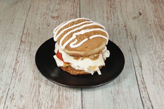 A burger with a sesame seed bun is placed on a black plate. The burger is topped with white icing-like drizzle, with visible ingredients like tomato slices and what appears to be layers of cream or cheese inside.