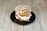 A burger with a sesame seed bun is placed on a black plate. The burger is topped with white icing-like drizzle, with visible ingredients like tomato slices and what appears to be layers of cream or cheese inside.