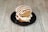 A burger with a sesame seed bun is placed on a black plate. The burger is topped with white icing-like drizzle, with visible ingredients like tomato slices and what appears to be layers of cream or cheese inside.