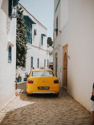 A narrow cobblestone street lined with white buildings featuring blue window frames and shutters. A yellow taxi is parked in the alley. There are some potted plants and vines climbing the walls, creating a picturesque, Mediterranean setting.