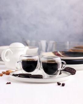 Two glass cups filled with espresso are placed on a white ceramic tray next to chocolate sandwich cookies and almonds. In the background, there are blurred kitchen items, including a white teapot and stacked plates.