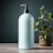 A sleek, light blue pump bottle stands prominently on a wooden surface. Beside it, a potted plant with lush green leaves provides a natural contrast. The background is a muted teal, enhancing the calm and clean aesthetic.