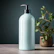 A sleek bottle of room spray on a wooden shelf surrounded by green plants.