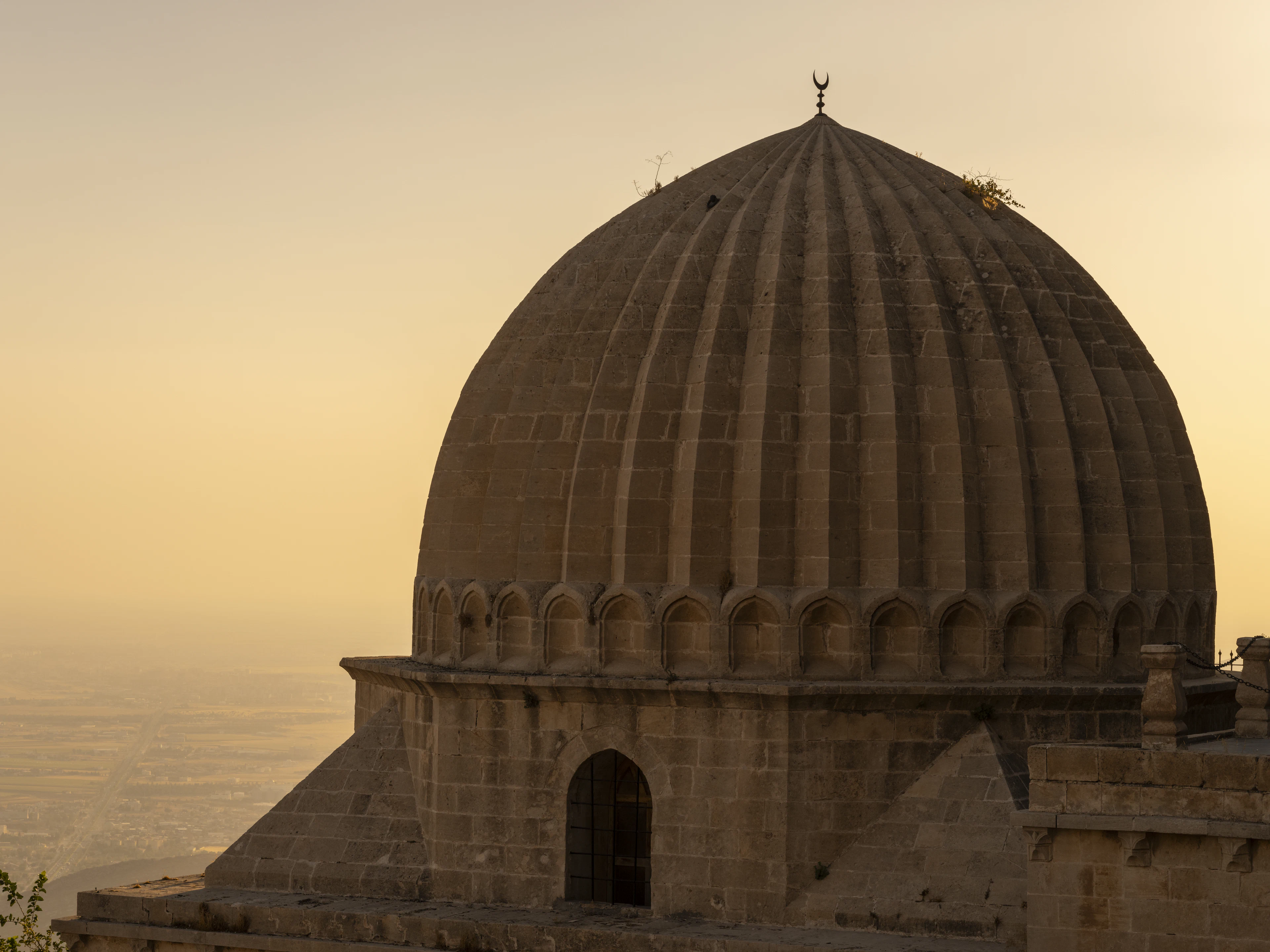 The fluted stone dome of a madrasa in Mardin in evening light, with the Mesopotamian plain fading into haze beyond