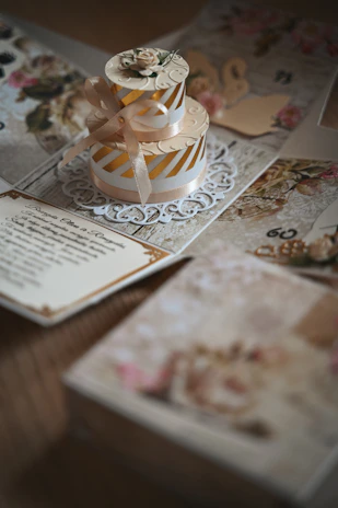 A beautifully designed card featuring a three-dimensional cake decoration with intricate floral and ribbon details. The background is adorned with floral patterns and elegant text, creating a vintage aesthetic.