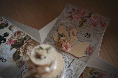 An intricately designed card featuring floral patterns, cursive handwriting, and decorative paper cutouts including a swan and pink roses. The card is open, revealing a layered structure with vintage elements and delicate details on each panel.