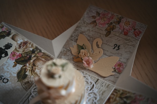 An intricately designed card featuring floral patterns, cursive handwriting, and decorative paper cutouts including a swan and pink roses. The card is open, revealing a layered structure with vintage elements and delicate details on each panel.