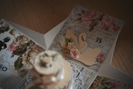 An intricately designed card featuring floral patterns, cursive handwriting, and decorative paper cutouts including a swan and pink roses. The card is open, revealing a layered structure with vintage elements and delicate details on each panel.