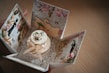 A beautifully crafted card box with an intricate design, featuring floral patterns and silhouettes of elegant figures. The center showcases a small decorative cake adorned with ribbons and a flower, surrounded by a lace-like embellishment.