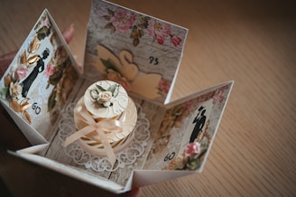 A beautifully crafted card box with an intricate design, featuring floral patterns and silhouettes of elegant figures. The center showcases a small decorative cake adorned with ribbons and a flower, surrounded by a lace-like embellishment.