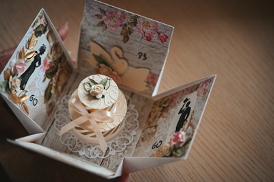 A beautifully crafted card box with an intricate design, featuring floral patterns and silhouettes of elegant figures. The center showcases a small decorative cake adorned with ribbons and a flower, surrounded by a lace-like embellishment.