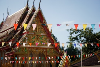An ornate temple with intricate details and vibrant colors is adorned with rows of colorful triangular flags strung across the foreground. The temple showcases traditional architectural motifs with a steeply pitched roof and elaborate carvings. The background consists of trees under a clear blue sky.