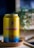 A yellow can of Athletic Brewing Co's Upside Dawn non-alcoholic brew is placed on a round wooden coaster. The can features a graphic design with blue mountains and an orange sun. In the background, there is a blurred green plant.