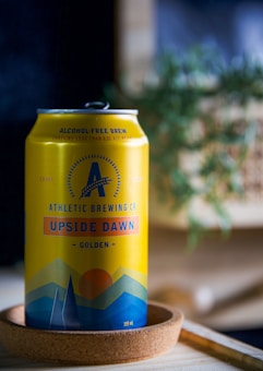 A yellow can of Athletic Brewing Co's Upside Dawn non-alcoholic brew is placed on a round wooden coaster. The can features a graphic design with blue mountains and an orange sun. In the background, there is a blurred green plant.
