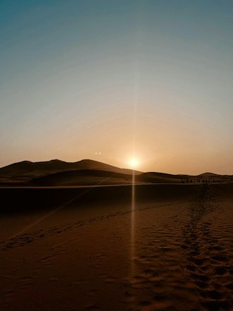 A vast desert landscape is illuminated by the setting sun, casting a golden glow over the sand dunes. The sun is low on the horizon, creating long shadows and highlighting the texture of the sand. A faint path of footprints leads into the distance, where a small caravan of camels is visible.