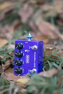 A purple guitar effects pedal labeled 'MXR Sub Machine' is nestled among grass and dried leaves. It features multiple knobs and switches with a prominent blue knob on top.