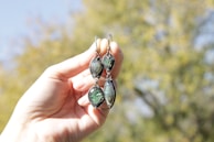 A hand holds two earrings with intricate designs featuring various stones. The earrings have teardrop shapes with a mix of green and iridescent stones. The background is blurred, showing hints of green foliage and blue sky, indicating an outdoor setting.