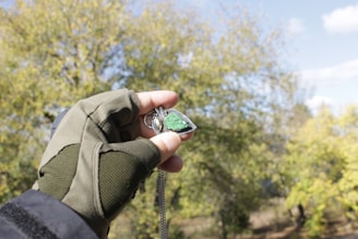 A hand wearing a green glove holds a necklace pendant with a green stone against a backdrop of trees and a partly cloudy sky. The pendant appears to be metallic and reflective, with a triangular shape.