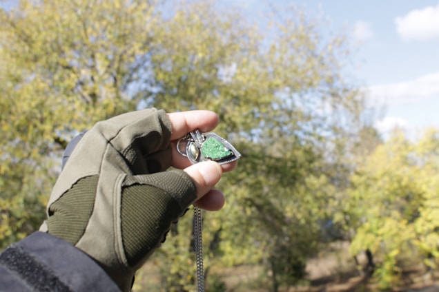 A hand wearing a green glove holds a necklace pendant with a green stone against a backdrop of trees and a partly cloudy sky. The pendant appears to be metallic and reflective, with a triangular shape.