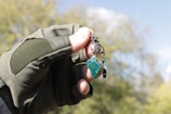 A hand wearing a tactical glove holds a keychain with a piece of green stone. The background consists of blurred greenery and a blue sky.