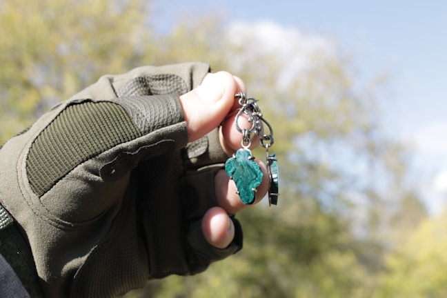 A hand wearing a tactical glove holds a keychain with a piece of green stone. The background consists of blurred greenery and a blue sky.