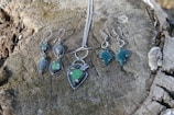 Several pieces of jewelry, including earrings and a necklace with green stones, are placed on a weathered tree stump. The jewelry has metal fittings, giving it a rustic and elegant look.