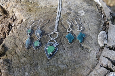 Several pieces of jewelry, including earrings and a necklace with green stones, are placed on a weathered tree stump. The jewelry has metal fittings, giving it a rustic and elegant look.