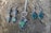A collection of silver jewelry is displayed on a textured, wooden surface. The set includes two pairs of dangling earrings and a pendant necklace. The earrings on the left feature elongated stones in green and iridescent gray, while the earrings on the right have uniquely shaped green stones. The necklace has a pendant with a matching green stone and silver accents, attached to a silver chain.