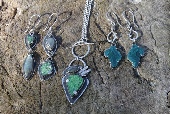A collection of silver jewelry is displayed on a textured, wooden surface. The set includes two pairs of dangling earrings and a pendant necklace. The earrings on the left feature elongated stones in green and iridescent gray, while the earrings on the right have uniquely shaped green stones. The necklace has a pendant with a matching green stone and silver accents, attached to a silver chain.