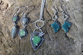 A collection of silver jewelry is displayed on a textured, wooden surface. The set includes two pairs of dangling earrings and a pendant necklace. The earrings on the left feature elongated stones in green and iridescent gray, while the earrings on the right have uniquely shaped green stones. The necklace has a pendant with a matching green stone and silver accents, attached to a silver chain.