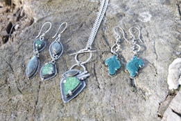 A collection of silver jewelry, including a pair of earrings and a necklace with geometric designs and green stones, is displayed on a textured wooden surface.