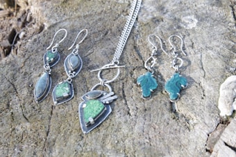 A collection of silver jewelry, including a pair of earrings and a necklace with geometric designs and green stones, is displayed on a textured wooden surface.