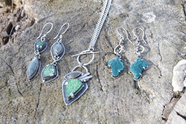 A collection of silver jewelry, including a pair of earrings and a necklace with geometric designs and green stones, is displayed on a textured wooden surface.