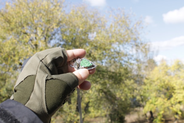 A hand wearing an olive-green glove holds a pendant with a shiny green gemstone against a backdrop of trees. The pendant hangs from a chain, and in the background, there are trees with autumn-colored leaves and a clear blue sky with scattered clouds.