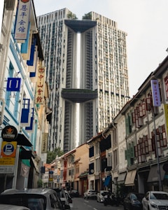 A modern high-rise building with a unique, symmetrical design featuring two towers connected by sky bridges stands prominently in the background. In the foreground, a narrow street is lined with traditional shophouses, showcasing colorful facades and wooden shutters. Banners with Chinese characters and advertisements add to the urban atmosphere. Several parked cars line the sides of the street, and a few people can be seen walking along the sidewalks.