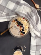 A bowl of oatmeal topped with banana slices, blackberries, nuts, and seeds is placed on a table. A white and gray checkered cloth is draped around it. Adjacent to the bowl, there is a wooden spoon with scattered oats.